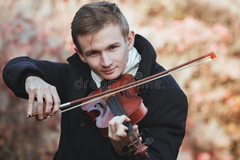 Portrait of a young elegant man playing the violin on autumn nature backgroung, a boy with a bowed orchestra instrument makes a concert, concept of classical music, hobby and art