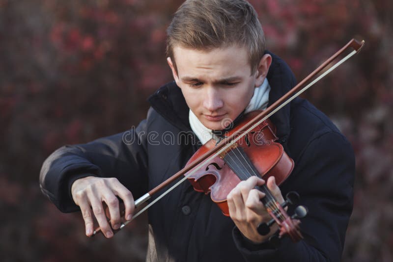 Portrait of a young elegant man playing the violin on autumn nature backgroung, a boy with a bowed orchestra instrument makes a concert, concept of classical music, hobby and art