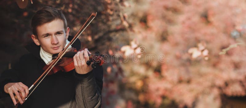 Portrait of a young elegant man playing the violin on autumn nature backgroung, a boy with a bowed orchestra instrument makes a concert, concept of classical music, hobby and art