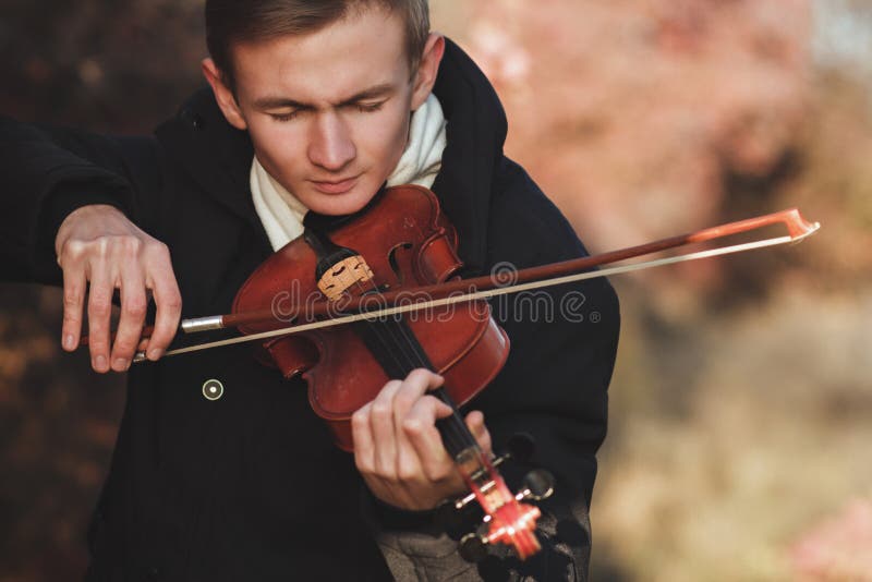 Portrait of a young elegant man playing the violin on autumn nature backgroung, a boy with a bowed orchestra instrument makes a concert, concept of classical music, hobby and art