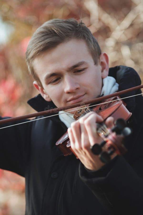 Portrait of a young elegant man playing the violin on autumn nature backgroung, a boy with a bowed orchestra instrument makes a concert, concept of classical music, hobby and art