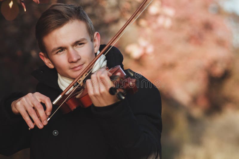 Portrait of a young elegant man playing the violin on autumn nature backgroung, a boy with a bowed orchestra instrument makes a concert, concept of classical music, hobby and art