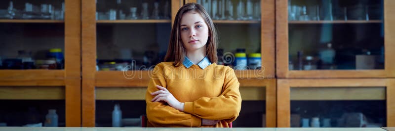 Portrait of young female college student in chemistry class, sitting behind the desk with crossed arms.