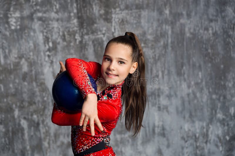 A portrait of young girl gymnast in a red suit makes exercise with a ball against a gray wall. She holds the ball