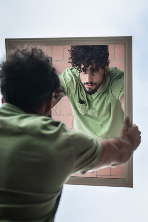 Portrait of a young man with beard and curly hair, who looks at himself while holding a mirror in his hands