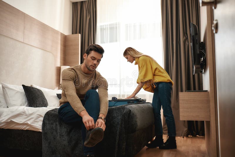 Portrait of young men and women in casual wear being together in their room in a hotel. Woman unpacks their luggage while men takes off his shoes. Travelling together concept. Horizontal shot. Portrait of young men and women in casual wear being together in their room in a hotel. Woman unpacks their luggage while men takes off his shoes. Travelling together concept. Horizontal shot