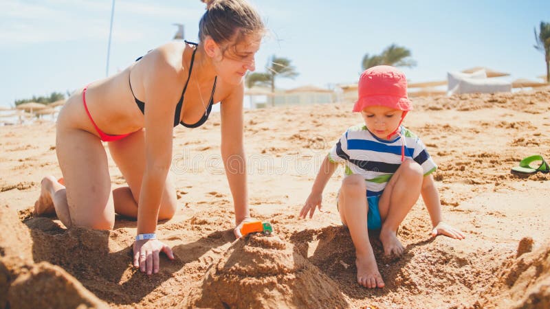 Portrait of young mother teaching her 3 years old toddler son building castles from sand on the sea beach. Family