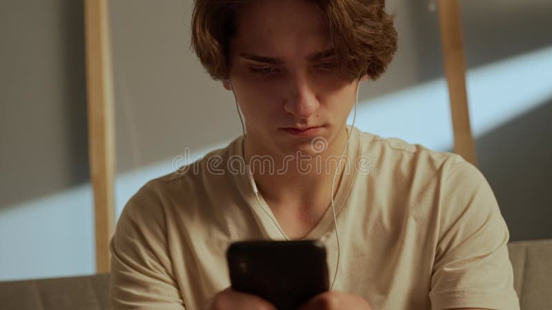 Portrait of young person in the room sitting on the couch. Close up of man face in earphones with smartphone, typing