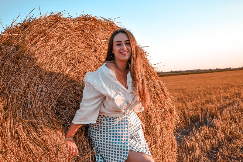 Portrait of young sexy woman on the haystack in morning sunlight, countryside. Beautiful woman in a dress sits on a haystack.