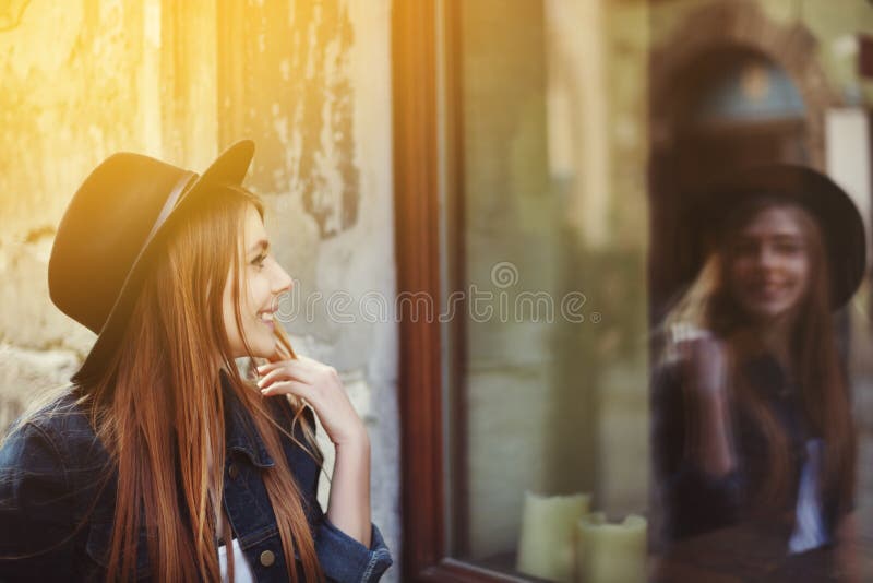 Portrait of young smiling woman looking at the shop window. Model wearing stylish wide-brimmed black hat. City lifestyle