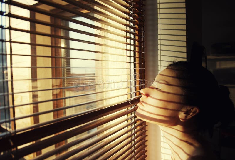 Portrait of young woman looking through sun blinds window shutter