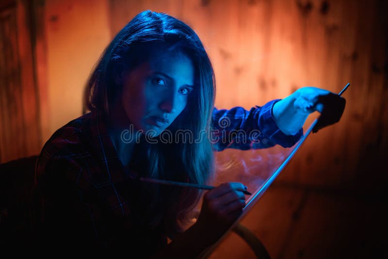 Portrait of young woman in room with blue lighting who paints on canvas with brush and looks at the camera