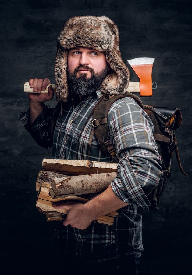 Portret brodaty woodcutter z plecakiem ubierał w kapeluszowej mienie łupce, ax szkocka krata trapera i koszula i Pracowniana fotografia przeciw zmrok textured ścianie