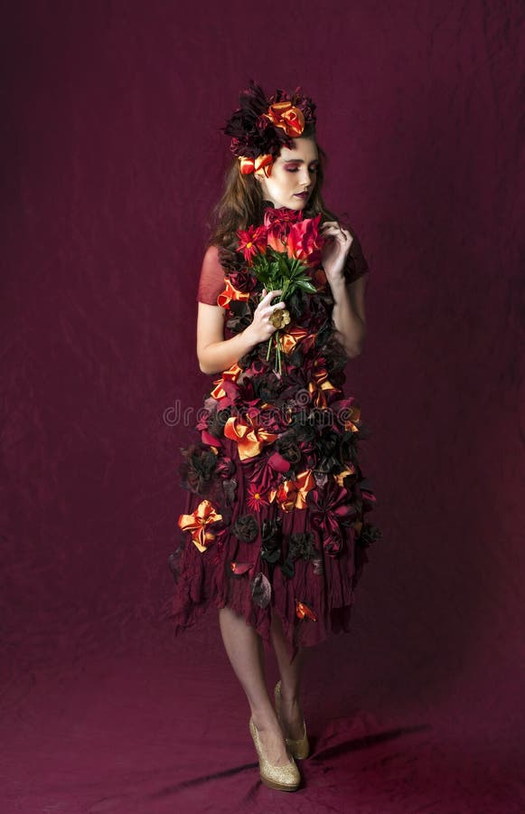 Color portrait of a beautiful brunette woman wearing a floral fantasy outfit of autumn colors posing in front of a red background, holding a bouquet of red flowers. Color portrait of a beautiful brunette woman wearing a floral fantasy outfit of autumn colors posing in front of a red background, holding a bouquet of red flowers