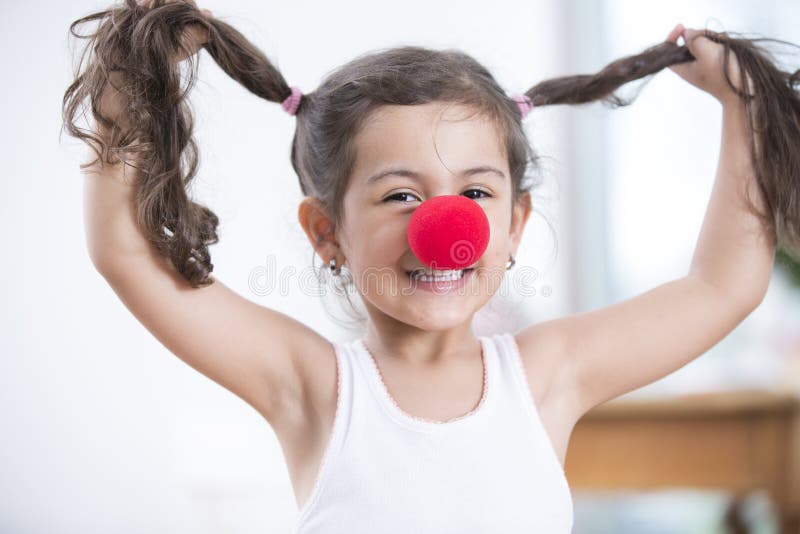 Portret jest ubranym błazenu nosa mienia pigtails w domu figlarnie mała dziewczynka zdjęcie stock Portret jest ubranym błazenu nosa mienia pigtails w domu figlarnie mała dziewczynka zdjęcie stock