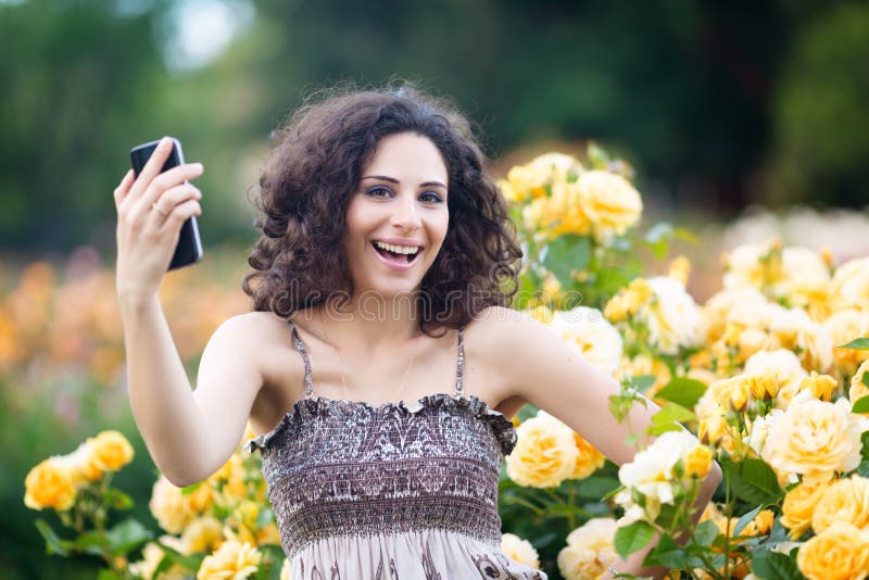 Portret Kaukaska kobieta z ciemnym kędzierzawym włosy bierze selfie blisko kolor żółty róży krzaków w ogródzie różanym, wideo wez obraz stock Portret Kaukaska kobieta z ciemnym kędzierzawym włosy bierze selfie blisko kolor żółty róży krzaków w ogródzie różanym, wideo wez obraz stock