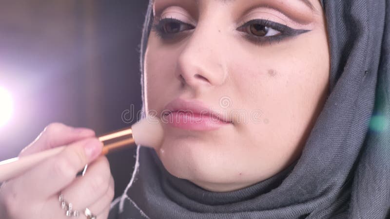 Portret kobieta wręcza robić makijażowi i stawiać highlighter dla młodej muzułmańskiej kobiety w hijab na jaskrawych światłach