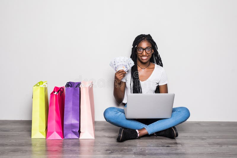 Portret młoda afro amerykańska kobieta używa laptop podczas gdy siedzący na podłodze z dolarów banknotami blisko toreb na zakupy obrazy stock Portret młoda afro amerykańska kobieta używa laptop podczas gdy siedzący na podłodze z dolarów banknotami blisko toreb na zakupy obrazy stock