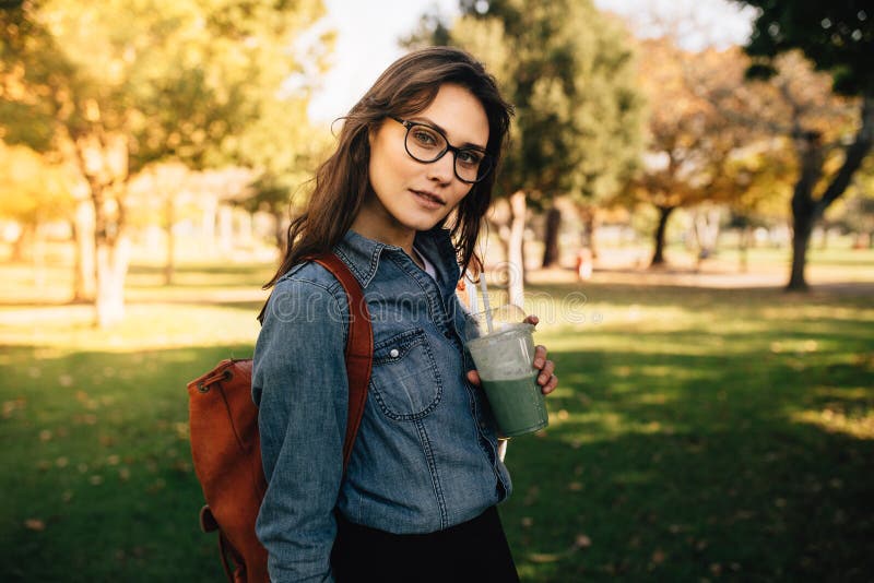 Portret piękna kobiety pozycja przy parkiem z owocowym sokiem Żeńska jest ubranym drelichowa koszula i eyeglasses z torby obwiesz obraz stock