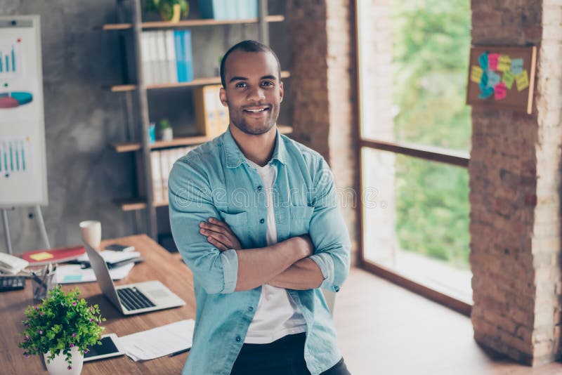 Portret pomyślny czarny facet, patrzejący kamerę, stoi z krzyżować rękami przy jego miejscem pracy w biurze w przypadkowym sma