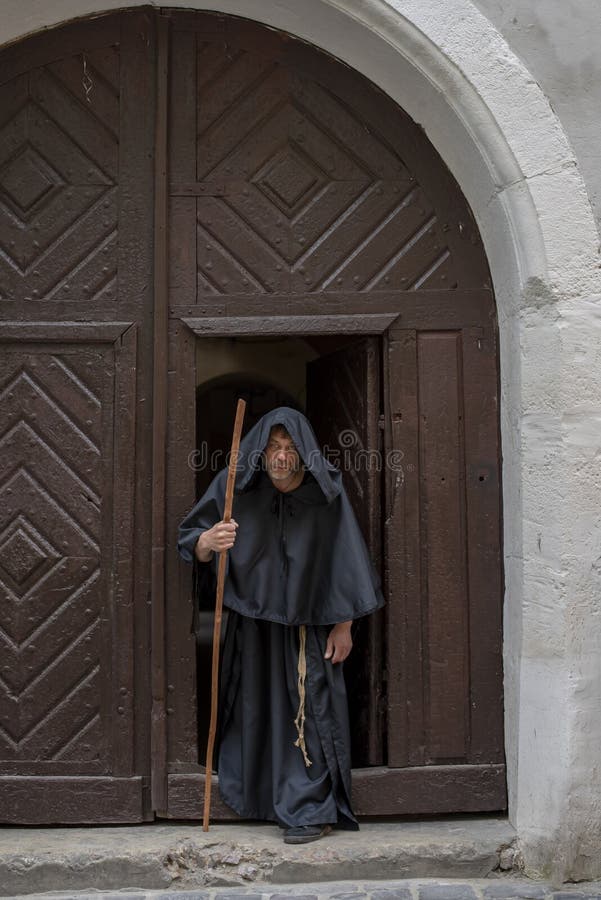 Portrait of an elderly hunched monk 45-50 years old with a beard in a black cassock with a staff coming out of the textured gate of a monastery or church, general plan. Portrait of an elderly hunched monk 45-50 years old with a beard in a black cassock with a staff coming out of the textured gate of a monastery or church, general plan.