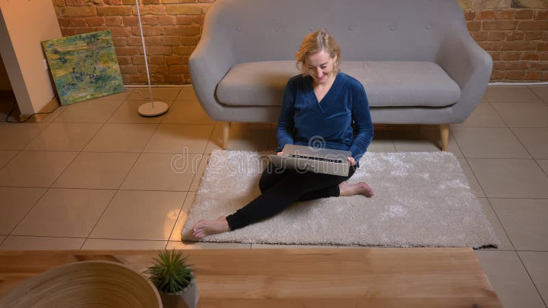 Portret starszy caucasian damy obsiadanie na podłodze i dopatrywanie w laptop szczęśliwie w wygodnej domowej atmosferze zdjęcie stock Portret starszy caucasian damy obsiadanie na podłodze i dopatrywanie w laptop szczęśliwie w wygodnej domowej atmosferze zdjęcie stock