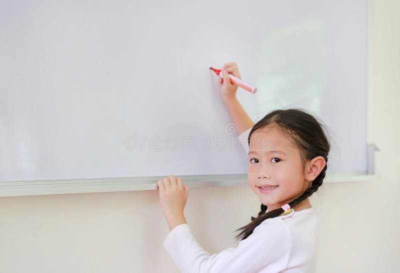 Portret szczęśliwa mała Azjatycka dziecko dziewczyna, uczennica pisze coś na whiteboard z markierem i patrzeje kamerę w lub obrazy royalty free Portret szczęśliwa mała Azjatycka dziecko dziewczyna, uczennica pisze coś na whiteboard z markierem i patrzeje kamerę w lub obrazy royalty free