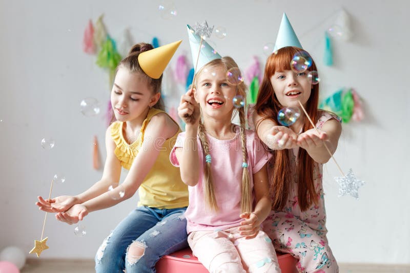 Portrait of three beautiful girls wear festive caps, play with bubbles, sit together on chair, celebrate birthday, being in good mood, use magic wand, have party in decorated room. Childhood concept. Portrait of three beautiful girls wear festive caps, play with bubbles, sit together on chair, celebrate birthday, being in good mood, use magic wand, have party in decorated room. Childhood concept