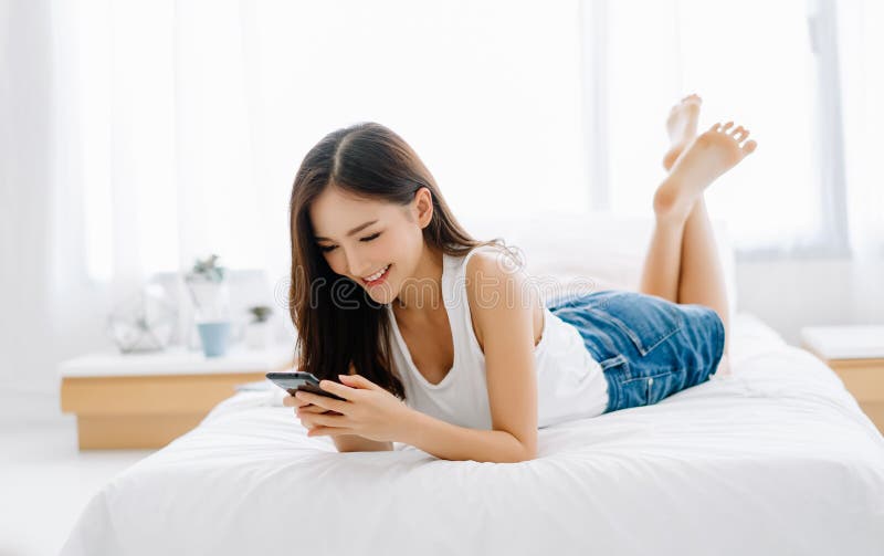 Portret uśmiechniętej pięknej Azjatki, czułej, świeżej, szczęśliwej twarzy leżącej na łóżku i wysyłającej SMS-y z smartfonem, jes zdjęcie royalty free Portret uśmiechniętej pięknej Azjatki, czułej, świeżej, szczęśliwej twarzy leżącej na łóżku i wysyłającej SMS-y z smartfonem, jes zdjęcie royalty free