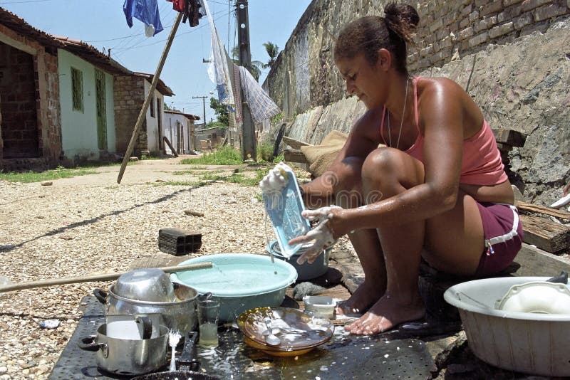 Brazil, state Pernambuco, Recife city: street from one of the most criminal slums of the city barrio of Santo Amaro. Poverty and unemployment in this slum is large and this woman is washing her utensils, cooking utensils, pots and pans in the tropical heat on the side of the road, but she has running water. Brazil, state Pernambuco, Recife city: street from one of the most criminal slums of the city barrio of Santo Amaro. Poverty and unemployment in this slum is large and this woman is washing her utensils, cooking utensils, pots and pans in the tropical heat on the side of the road, but she has running water.