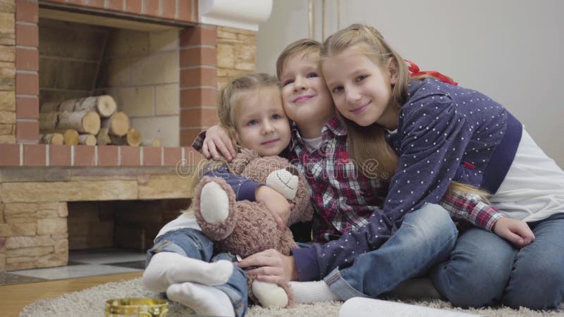 Portret van drie Kaukasische kinderen die naar camera kijken en glimlachen Goedaardige meisjes en jongen die thuis knuffelen voor