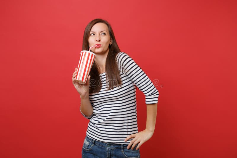 Portret van dromerige vrij jonge vrouw die in gestreepte kleren kola of soda van plastic die kop drinken op helder rood wordt geï stock fotografie Portret van dromerige vrij jonge vrouw die in gestreepte kleren kola of soda van plastic die kop drinken op helder rood wordt geï stock fotografie