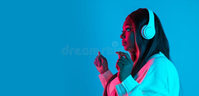 Music. Studio shot of African charming woman with long straight hair isolated on blue studio background in neon light. Concept of human emotions, facial expression, beauty, fashion. Bodypositive and diversity. Music. Studio shot of African charming woman with long straight hair isolated on blue studio background in neon light. Concept of human emotions, facial expression, beauty, fashion. Bodypositive and diversity