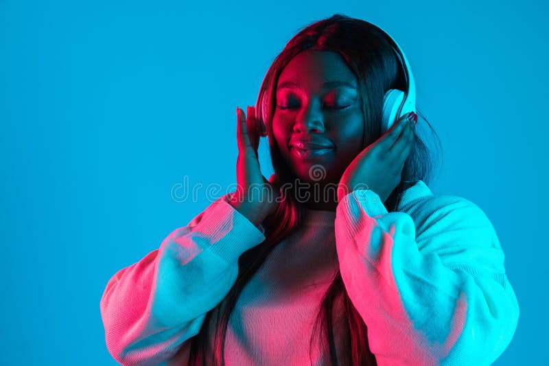 Music. Studio shot of African charming woman with long straight hair isolated on blue studio background in neon light. Concept of human emotions, facial expression, beauty, fashion. Bodypositive and diversity. Music. Studio shot of African charming woman with long straight hair isolated on blue studio background in neon light. Concept of human emotions, facial expression, beauty, fashion. Bodypositive and diversity