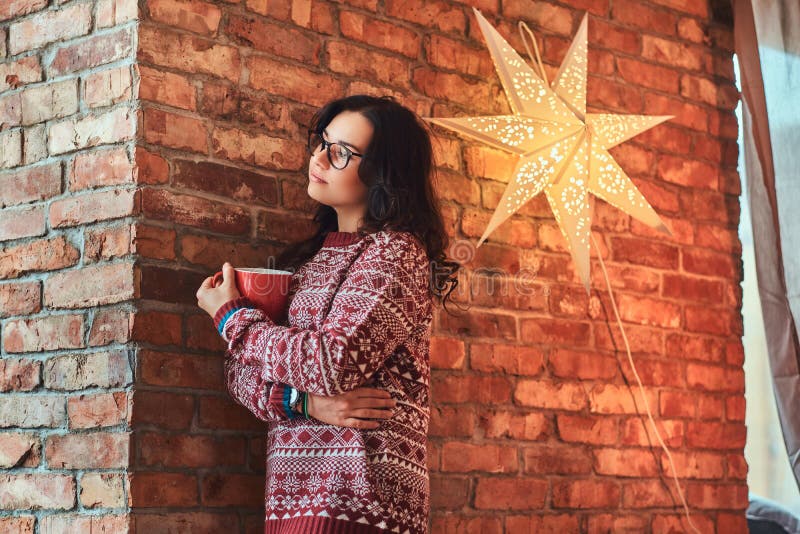 Portret van een eenzaam donkerbruin meisje die oogglazen en warme sweater dragen die een kop van koffie houden terwijl het leunen royalty-vrije stock afbeelding Portret van een eenzaam donkerbruin meisje die oogglazen en warme sweater dragen die een kop van koffie houden terwijl het leunen royalty-vrije stock afbeelding