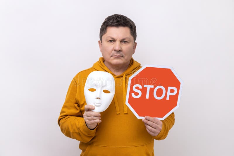 Portrait of serious dark haired man holding white mask with unknown face and red traffic sign, looking at camera, wearing urban style hoodie. Indoor studio shot isolated on white background. Portrait of serious dark haired man holding white mask with unknown face and red traffic sign, looking at camera, wearing urban style hoodie. Indoor studio shot isolated on white background.