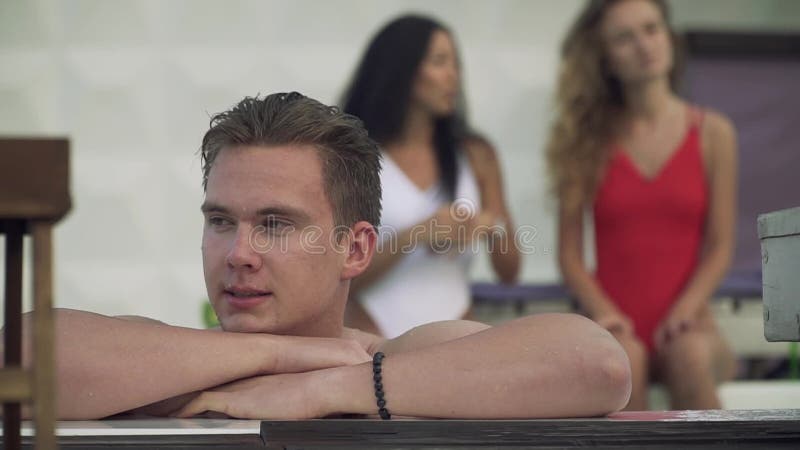 Portret van een glimlachende jonge mens in de pool op de achtergrond van twee mooie speelse meisjes in zwempakken Een bedrijf van