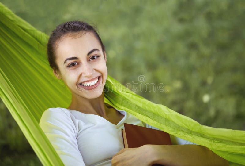 Portret van een glimlachende jonge vrouw die in groene hangmat met boek in park liggen royalty-vrije stock foto's Portret van een glimlachende jonge vrouw die in groene hangmat met boek in park liggen royalty-vrije stock foto's