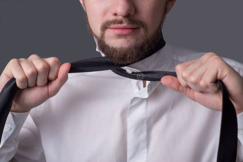 Portret van een jonge bebaarde man in een wit shirt die zichzelf probeert te wurgen met zijn eigen stropdas Op een donkergrijze a stock foto Portret van een jonge bebaarde man in een wit shirt die zichzelf probeert te wurgen met zijn eigen stropdas Op een donkergrijze a stock foto
