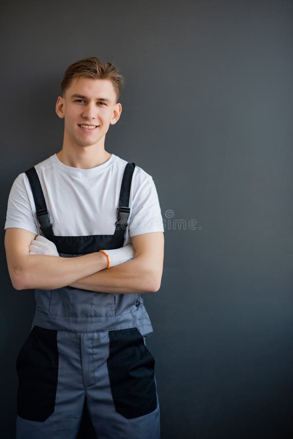 Portret van een jonge glimlachende beroepsarbeider in grijze overall en wit shirt dat staat met een kruis op een grijs stock foto Portret van een jonge glimlachende beroepsarbeider in grijze overall en wit shirt dat staat met een kruis op een grijs stock foto