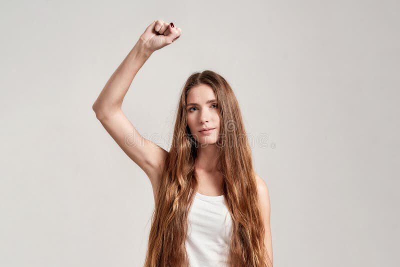 Portrait of young caucasian woman with long hair wearing white shirt looking at camera while showing, raising clenched fist, standing isolated over grey background. Front view. Horizontal shot. Portrait of young caucasian woman with long hair wearing white shirt looking at camera while showing, raising clenched fist, standing isolated over grey background. Front view. Horizontal shot