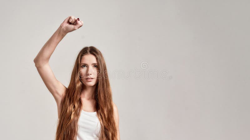 Portrait of young caucasian woman with long hair wearing white shirt having confident look while showing, raising clenched fist, standing  over grey background. Front view. Web Banner. Portrait of young caucasian woman with long hair wearing white shirt having confident look while showing, raising clenched fist, standing  over grey background. Front view. Web Banner