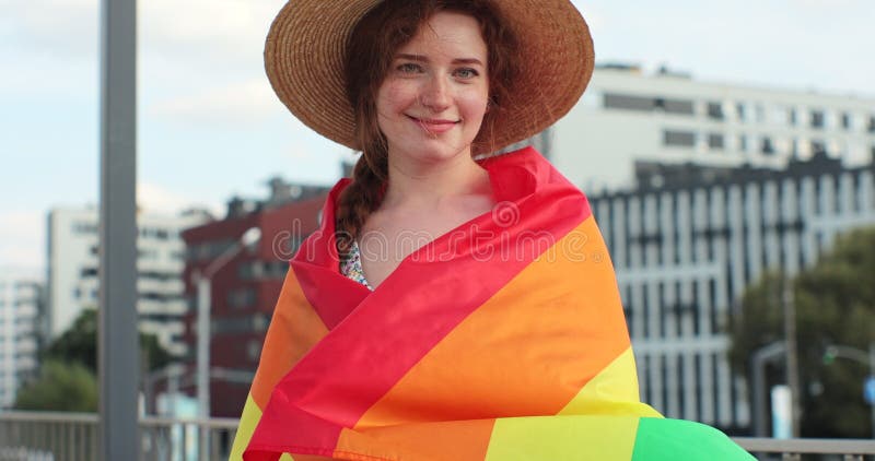 Portrait of a beautiful young woman in a hat with long red hair covering herself in lgbt flag while looking to camera. Alone. One. Keeping fist up, covering LGBT flag. Portrait of a beautiful young woman in a hat with long red hair covering herself in lgbt flag while looking to camera. Alone. One. Keeping fist up, covering LGBT flag