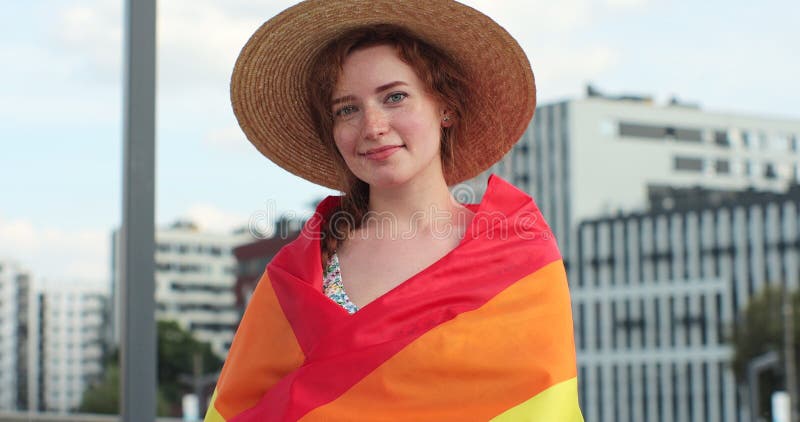 Portrait of a beautiful young woman in a hat with long red hair covering herself in lgbt flag while looking to camera. Alone. One. Keeping fist up, covering LGBT flag. Portrait of a beautiful young woman in a hat with long red hair covering herself in lgbt flag while looking to camera. Alone. One. Keeping fist up, covering LGBT flag