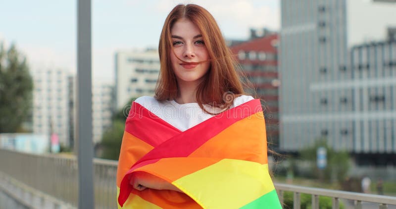 Portrait of a beautiful young woman with long red hair covering in a rainbow flag. Lesbian woman standing at city street. Portrait of a beautiful young woman with long red hair covering in a rainbow flag. Lesbian woman standing at city street.