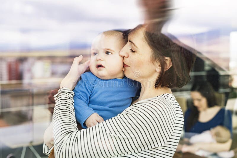 Portrait of new mom holding baby, standing by window in the cafe. Spending free time during maternity leave with friends in city. Portrait of new mom holding baby, standing by window in the cafe. Spending free time during maternity leave with friends in city.