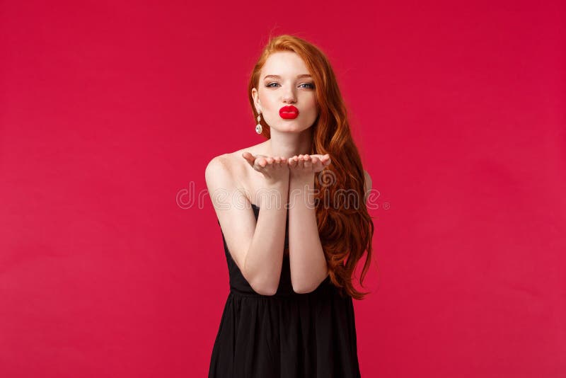 Portrait of coquettish feminine young beautiful redhead woman in black stylish dress, sending air kiss on palms near folded lips, looking at camera give sensual mwah, red background. Portrait of coquettish feminine young beautiful redhead woman in black stylish dress, sending air kiss on palms near folded lips, looking at camera give sensual mwah, red background.