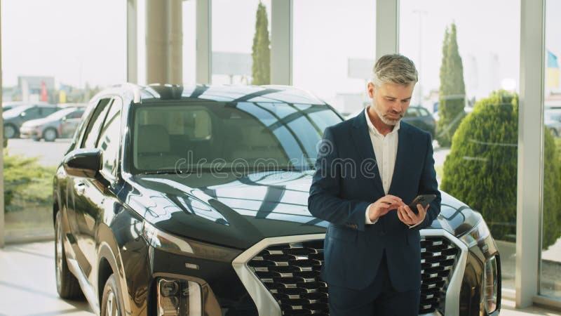 Portret van een rijpe , rijpe beard grijze haren man in een pak met smartphone die op de auto leunt en denkt of