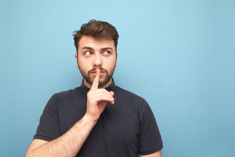 Portrait of an adult man with a beard thoughtfully looking sideways to a blank place and showing a sign of silence holding a finger near his mouth on a blue background. Isolated. Portrait of an adult man with a beard thoughtfully looking sideways to a blank place and showing a sign of silence holding a finger near his mouth on a blue background. Isolated