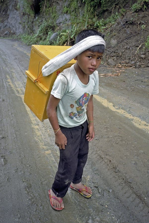 Philippines: Portrait of shy or frightened young ice cream vendor with his merchandise, ice in a cooler hanging at his head on a muddy road in the mountain village, Banaue on the island Luzon. He walks through the mud in flip flops with bare feet. Many Philippines are very poor and therefore every family member has to work to contribute sufficient money for everyday life, existence. Child labor in the Philippines is a widespread phenomenon and is a violation of the rights of the child, children. Philippines: Portrait of shy or frightened young ice cream vendor with his merchandise, ice in a cooler hanging at his head on a muddy road in the mountain village, Banaue on the island Luzon. He walks through the mud in flip flops with bare feet. Many Philippines are very poor and therefore every family member has to work to contribute sufficient money for everyday life, existence. Child labor in the Philippines is a widespread phenomenon and is a violation of the rights of the child, children.